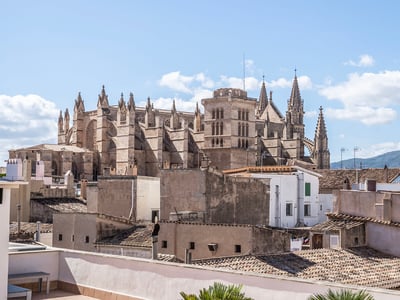 Una gran catedral gótica se alza sobre los tejados de terracota de una ciudad mediterránea bajo un cielo azul.