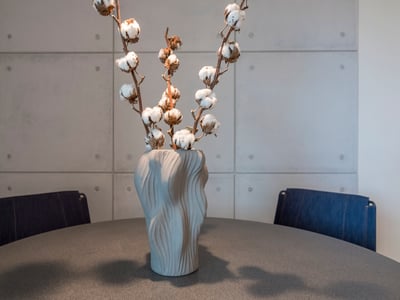 Jarrón gris texturizado con ramas de algodón sobre una mesa oscura frente a una pared de paneles de hormigón.