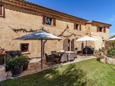 Sunny stone terrace with a dining table, wicker chairs, and white umbrellas next to a green lawn.