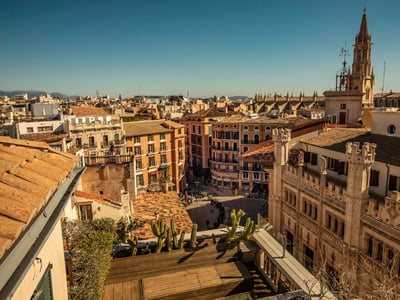 Erhöhte Ansicht über mediterrane Hausdächer und die Spitze einer gotischen Kathedrale unter strahlend blauem Himmel.