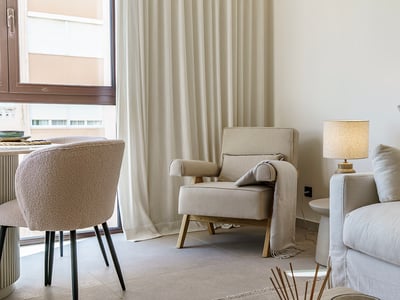 Rincón de salón luminoso con sofá crema, sillón, silla de lana rizada y cortinas suaves de suelo a techo.