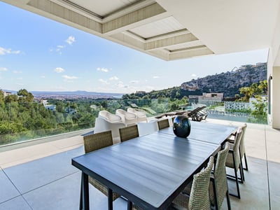 Terrasse moderne avec table à manger et garde-corps en verre surplombant une forêt et la ville au loin.