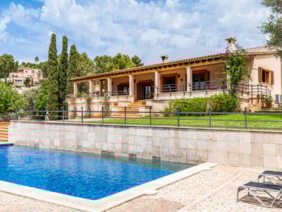 Gran piscina azul y tumbonas frente a una villa mediterránea con muro de piedra, jardín verde y terraza cubierta.