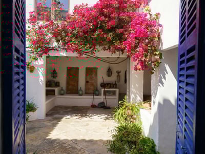 A sun-drenched white courtyard framed by blue shutters, featuring vibrant pink bougainvillea and rustic stone flooring.