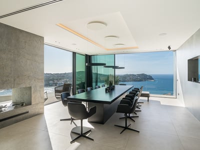 A modern dining room with a long black table, stone fireplace, and floor-to-ceiling windows overlooking the ocean.