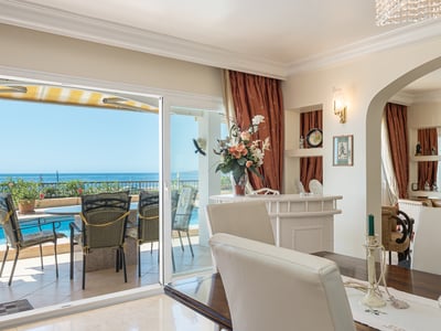 Interior dining area with a wooden table and white chairs looking out to a terrace with a pool and sea views.