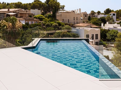 Infinity-Pool mit blauen Mosaikfliesen und Glasgeländer mit Blick auf eine grüne mediterrane Stadt.