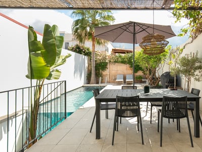 Modern outdoor terrace with a black dining table under an umbrella, next to a turquoise swimming pool and palm trees.