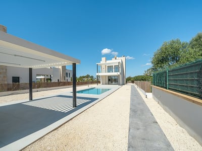 Exterior of a modern villa featuring a blue rectangular pool, white pergola, and gravel courtyard under a clear sky.
