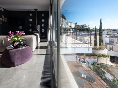 Modern living room with polished concrete floor, beige sofa, purple ottoman, and views of a sunny terrace through large glass doors.