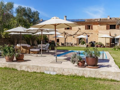 Large blue swimming pool surrounded by a stone deck with white umbrellas and wicker lounge furniture in a sunny garden.