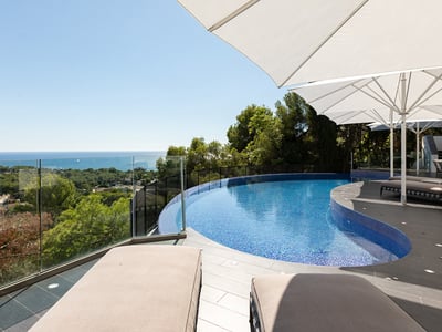 A curved blue swimming pool on a modern stone terrace with white umbrellas and sunbeds overlooking the sea.