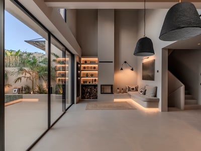 Minimalistiskt vardagsrum med högt i tak, inbyggd soffa, öppen spis och stora glasdörrar mot en tropisk uteplats.