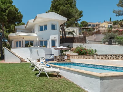 White two-story villa with a swimming pool, loungers, and green lawn surrounded by tall pine trees.