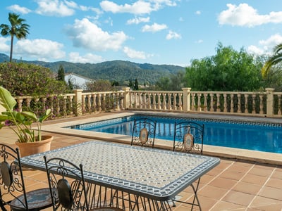Piscina rectangular y mesa de mosaico en una terraza de terracota con vistas a la montaña y palmeras.