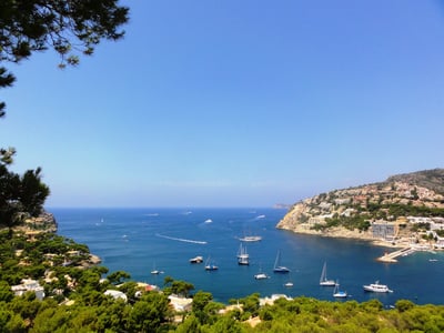 High-angle view of a blue bay with sailboats, surrounded by green hills and Mediterranean villas under a clear sky.