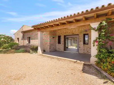 Entré till en stenvilla med träveranda, grusgång och blommande växter under en blå himmel.