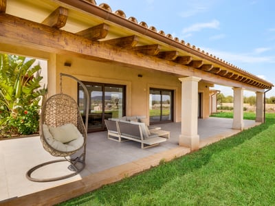 Övertäckt terrass med träbjälkar, moderna möbler, hängstol och gräsmatta under blå himmel.