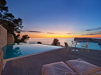 Infinitypool på trädäck med utsikt över havet vid solnedgången, utemöbler och glasräcke.