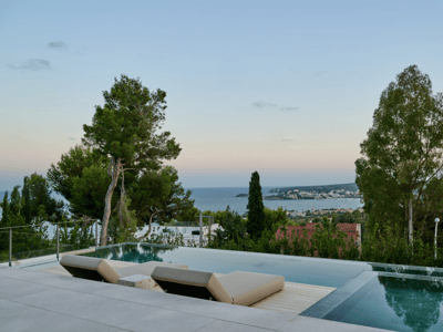 Piscina infinita moderna con dos tumbonas beige frente al mar azul y una ciudad costera al atardecer.