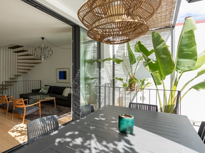Modern living room with a floating wooden staircase seen through open glass doors from a terrace with tropical plants.