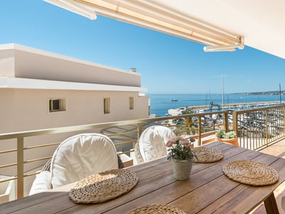 Balcón con mesa de madera y sillas blancas con vistas panorámicas a un puerto deportivo y al mar azul.