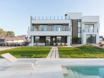 Villa moderna blanca de dos plantas con piscina turquesa, tumbona y césped bajo un cielo despejado.