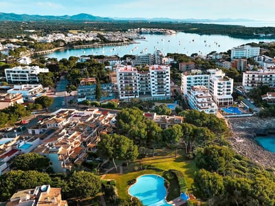 Drönarvy över en medelhavsstad med vita lägenhetshus, pooler och en blå vik med båtar.