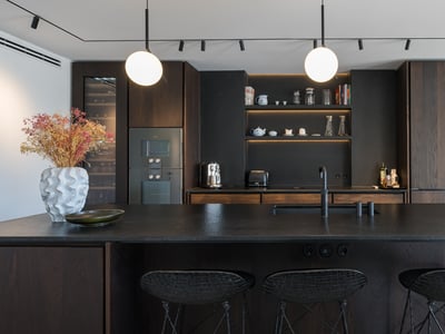 Cocina moderna con armarios de madera oscura, isla negra texturizada con taburetes y lámparas colgantes esféricas.