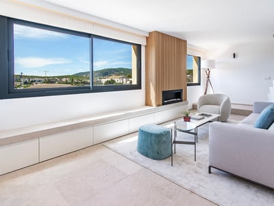 Salón luminoso con sofá gris, otomana turquesa, chimenea de listones de madera y gran ventanal con vistas.