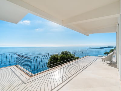 Bred modern terrass med stengolv och svart räcke med utsikt över ett lugnt blått hav under en klar himmel.