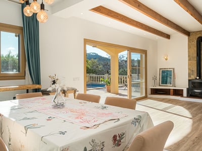 Comedor luminoso con vigas de madera, chimenea de piedra y puertas de cristal hacia una terraza con piscina.