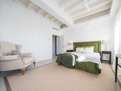 Helles Schlafzimmer mit weißen Balken, grünem Samtbett, beige Sessel und großen Fenstern.