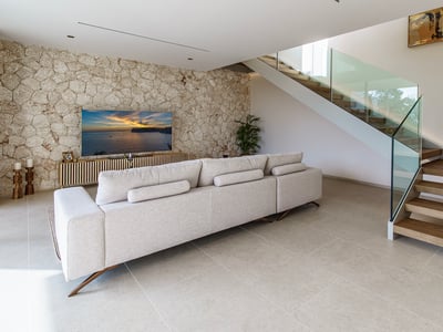 Salón amplio con sofá beige, pared de piedra decorativa, televisión y una escalera de madera con barandilla de vidrio.