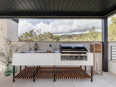 Moderne Außenküche mit Profi-Grill, Spüle und weißen Schränken auf einer Terrasse mit Blick auf die Berglandschaft.