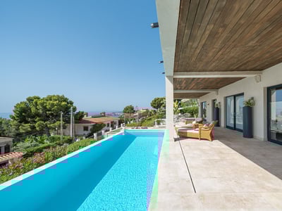 A bright terrace with a turquoise infinity pool, wicker loungers, and a wooden ceiling overlooking a coastal landscape.