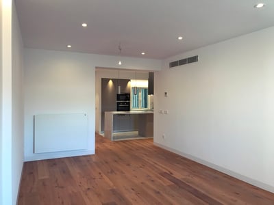 Geräumiges, leeres Wohnzimmer mit Parkettboden, das in eine moderne graue Küche mit Deckeneinbauleuchten führt.