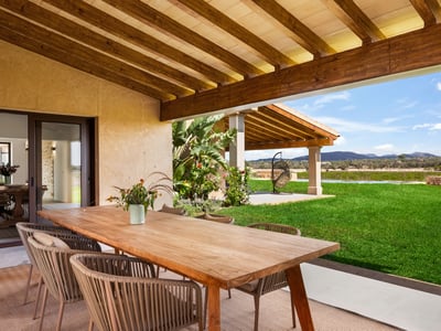 Övertäckt terrass med stort träbord och designstolar med utsikt över grön gräsmatta och kullar.