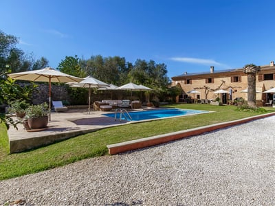View of a large stone villa with a blue swimming pool, white umbrellas, and garden under a clear sky.