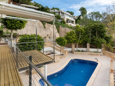 Vista en ángulo de una piscina azul con terraza de madera, patio de baldosas y vegetación en una colina.