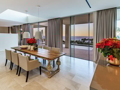 Elegant dining area featuring a long wooden table and floor-to-ceiling windows overlooking the ocean at sunset.