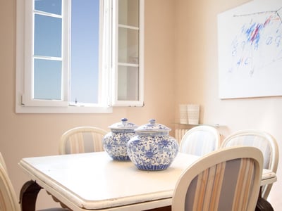 Comedor luminoso con mesa blanca, dos jarrones de porcelana azul y blanca y ventana abierta al cielo azul.