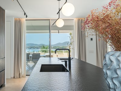 Cocina moderna con isla de piedra oscura y fregadero negro con vistas a una terraza soleada y al mar.