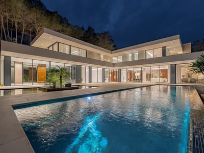 A luxury swimming pool in front of a glowing modern glass villa under a dark night sky.