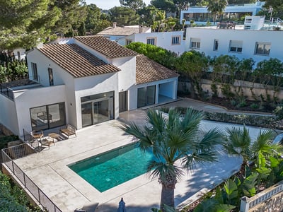 Aerial view of a white villa with a tiled roof, turquoise swimming pool, and large stone terrace surrounded by pine and palm trees.