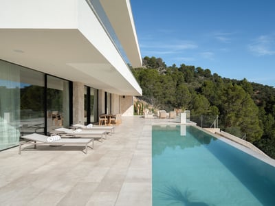 A spacious white stone terrace featuring an infinity pool, sun loungers, and views of a pine forest under a blue sky.