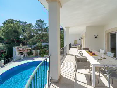 Övertäckt vit terrass med ett långt matbord och utsikt över en blå pool och lummig grönska.