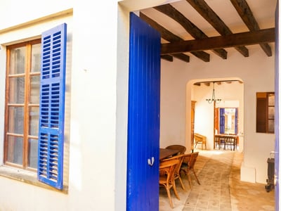 White exterior wall with bright blue shutters and open door leading to a rustic interior with wooden beams.