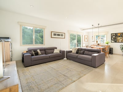 Rymligt och ljust vardagsrum med två grå soffor, ljusa klinkergolv och matplats i bakgrunden med stora fönster.