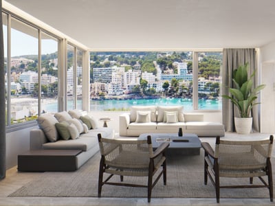 Bright living room with cream sofas and woven wood chairs facing a large window overlooking a Mediterranean bay.